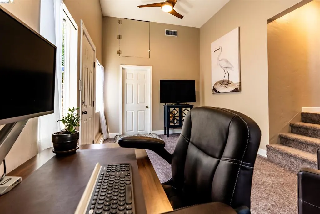 The spacious bonus room showcases a vaulted ceiling with a ceiling fan, neutral wall tones, carpeted flooring, an interior door with paneling to the laundry room, a separate door to the side yard patio, and a staircase to the kitchen