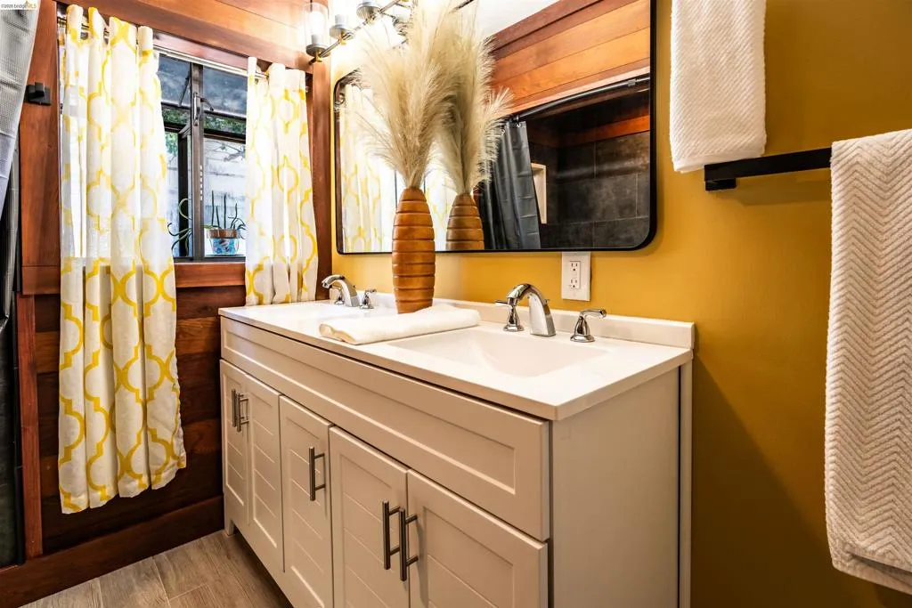 Dual vanity with white countertop and integrated sinks, matte black framed mirror, and teak wood-paneled wall accents. A charming bay window fills the room with soft light, providing views of the backyard