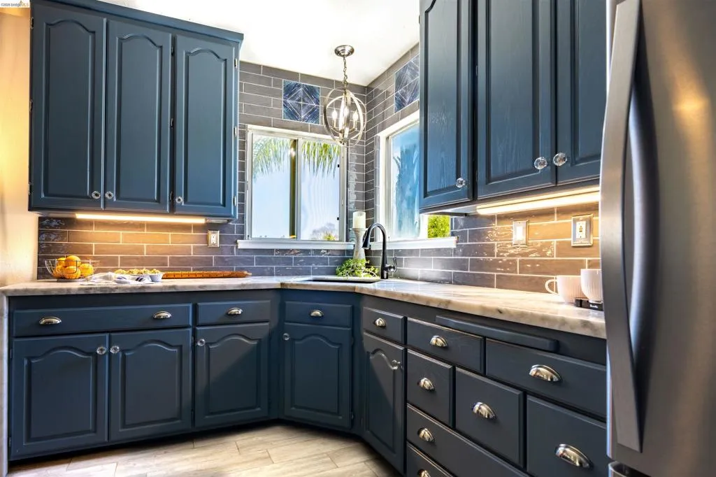Kitchen featuring blue cabinetry, light-toned countertops, a dark subway tile backsplash, stainless steel appliances, and under-cabinet lighting