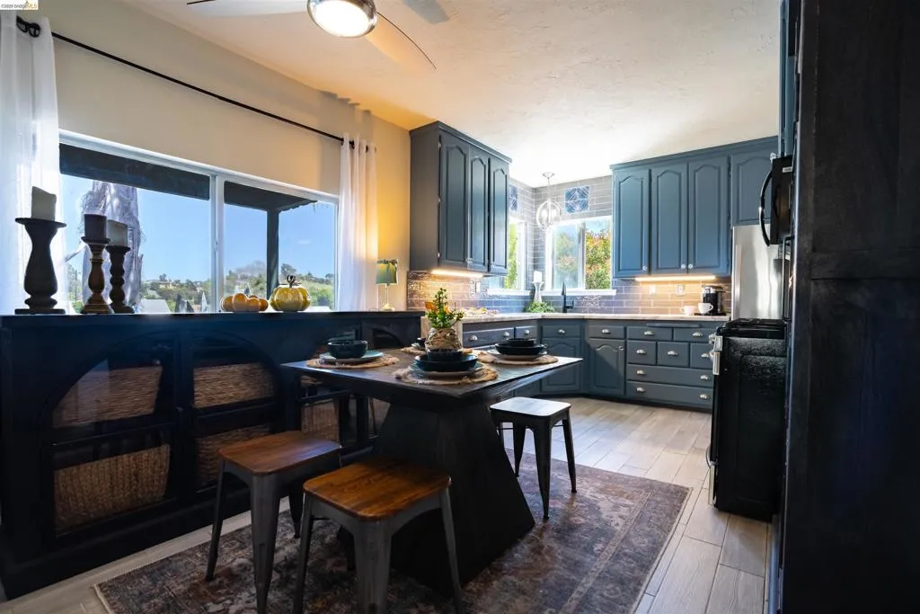 Kitchen featuring blue cabinetry, light wood-finish flooring, a tiled backsplash, built-in under-cabinet lighting, and a stainless-steel refrigerator. Each window frame offers stunning panoramic views of the Vallejo hills