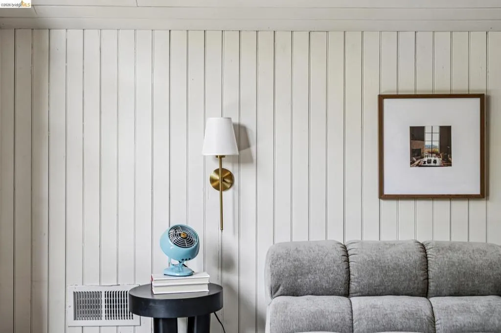Vertical white paneled wall featuring a brass-finish wall sconce with a white fabric shade and a floor vent