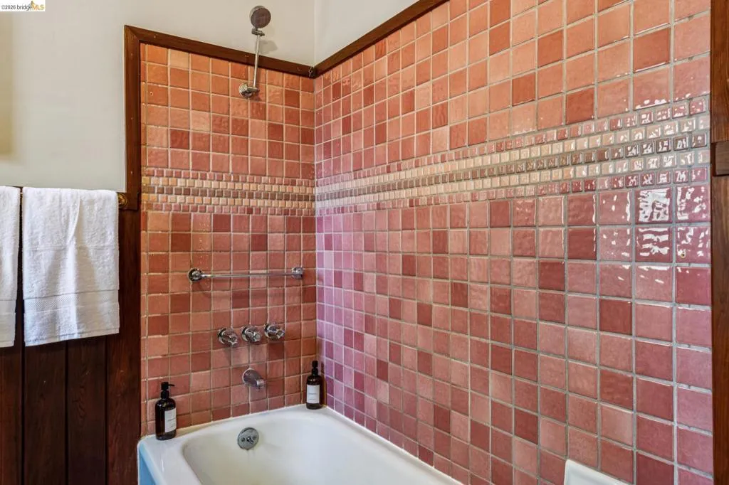 Bathtub and shower combination featuring a tiled surround in varying shades of pink and red, accented by a decorative mosaic tile band