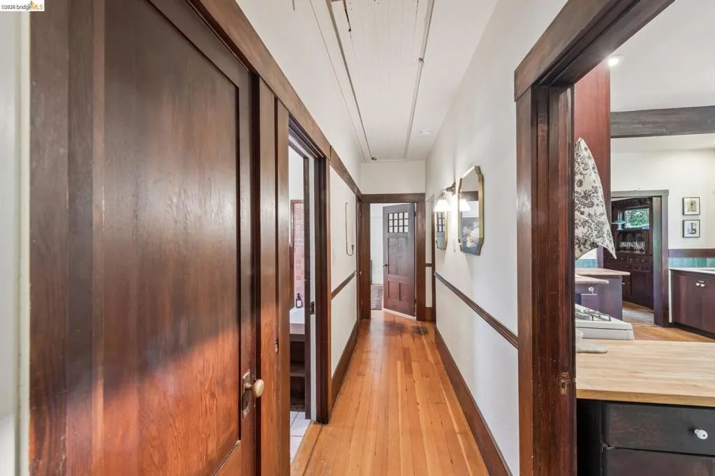 Hardwood flooring throughout a hallway with dark wood trim and doors, featuring wall-mounted sconces and a glimpse into a kitchen with light wood countertops