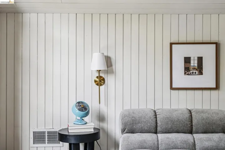 Vertical white paneled wall featuring a brass-finish wall sconce with a white fabric shade and a floor vent