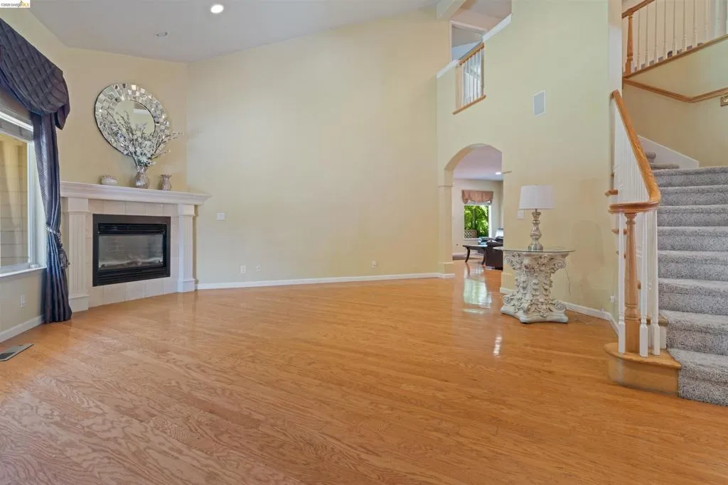 Spacious living area featuring wood-finish flooring, a fireplace with a mantel, high ceilings, an elegant staircase with carpeted treads and wood railings, and an arched doorway