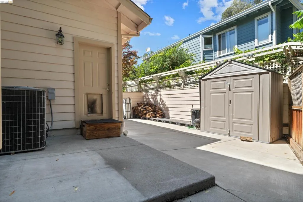 Concrete patio space with a side door entry, featuring an exterior light fixture, a compact storage shed, and a privacy fence with lattice detailing