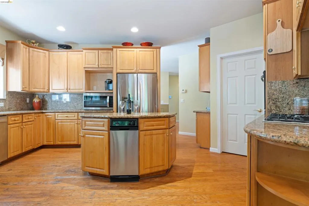 Kitchen featuring a central island with integrated dishwasher, wood-finish flooring, light wood cabinetry, stone countertops, and stainless steel appliances
