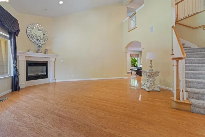 Spacious living area featuring wood-finish flooring, a fireplace with a mantel, high ceilings, an elegant staircase with carpeted treads and wood railings, and an arched doorway