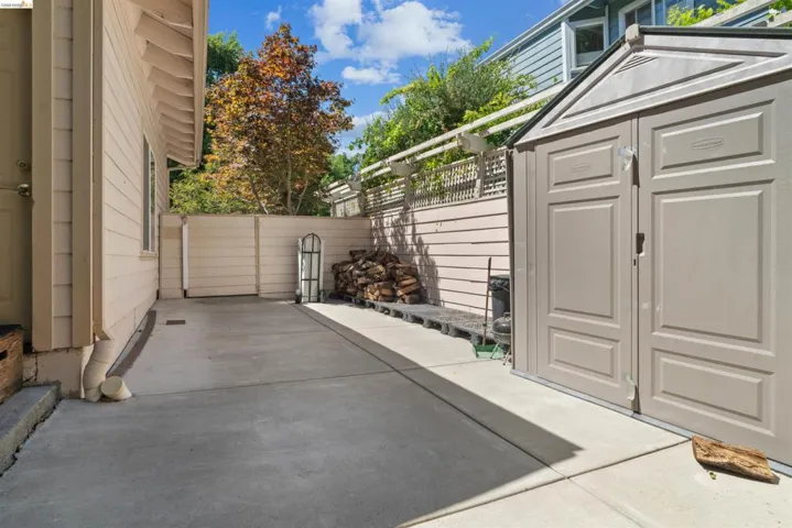 Concrete patio area featuring a beige storage shed with double doors, wood-paneled fencing, and a lattice-top privacy screen