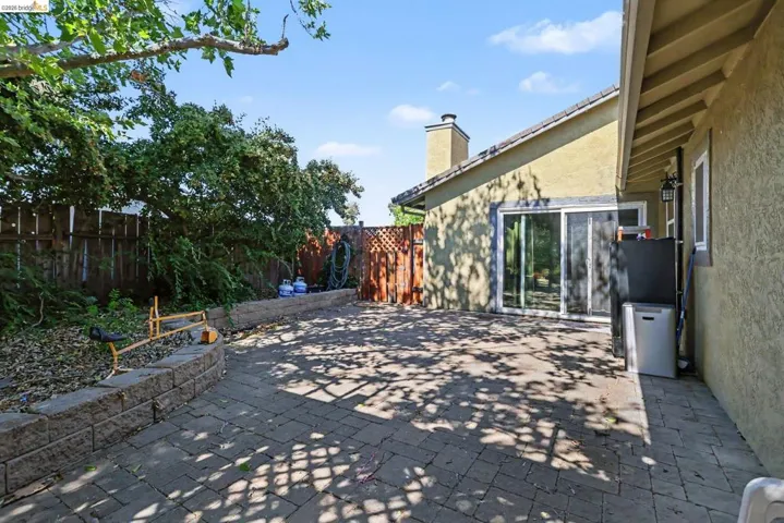 Spacious paved patio area featuring a retaining wall, mature perimeter foliage, and direct access to the interior via a sliding glass door