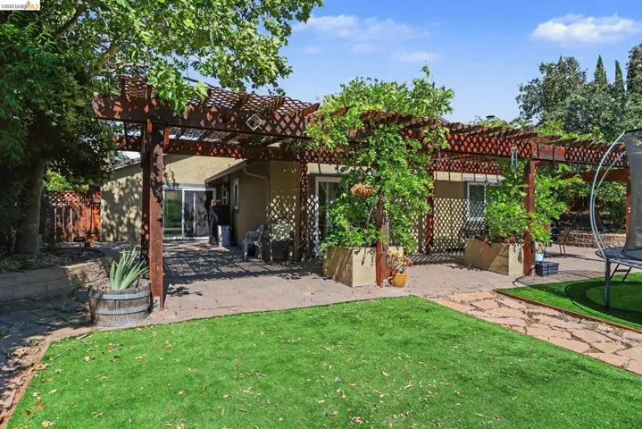 Expansive outdoor living area featuring a large wood-finish pergola, a stone paver patio, lush green lawn, mature trees, and a sliding glass door entry to the interior
