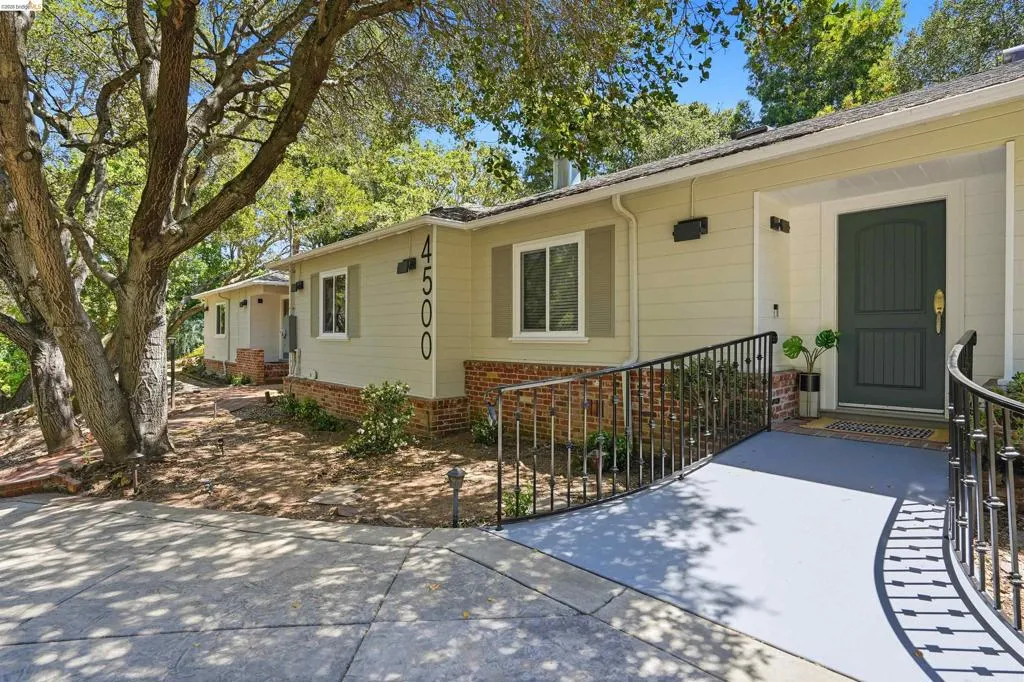 Exterior featuring light-toned siding, brick detailing, a dark green entry door, a curved entry ramp with black metal railings, and mature trees providing natural shade