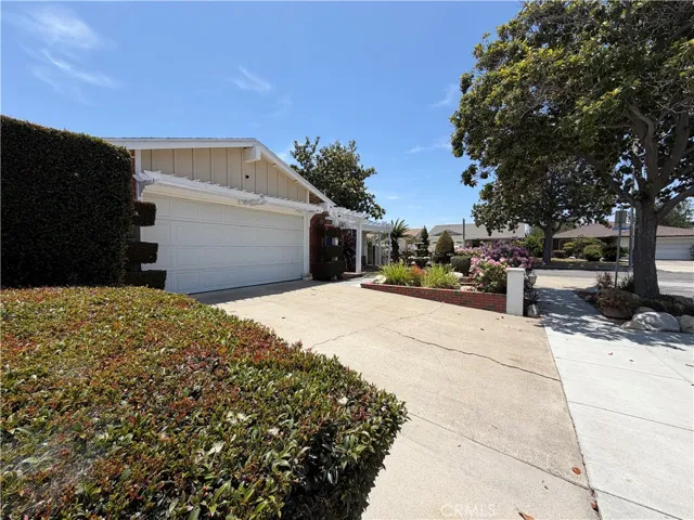 Charming front exterior with mature landscaping, a welcoming pergola entry, spacious driveway, attached garage, and a beautifully shaded sidewalk.