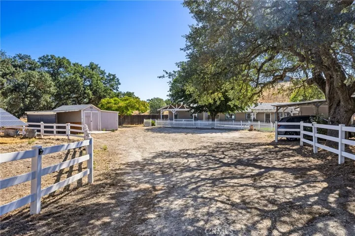  As you enter the property, a wide driveway framed by white rail fencing, mature oak trees, and open acreage leads to the main residence, guest house, and outbuildings, showcasing the property's peaceful country setting.