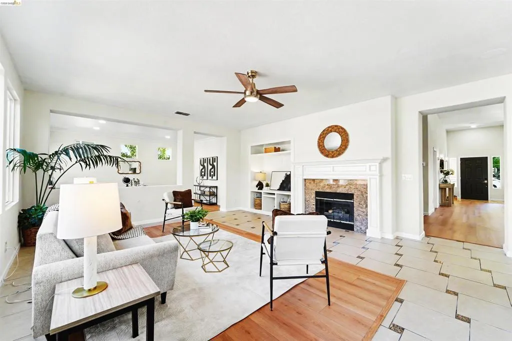 Spacious living area featuring a fireplace with a stone surround and white mantel, built-in shelving, a ceiling fan, and mixed flooring with wood-finish and tile elements