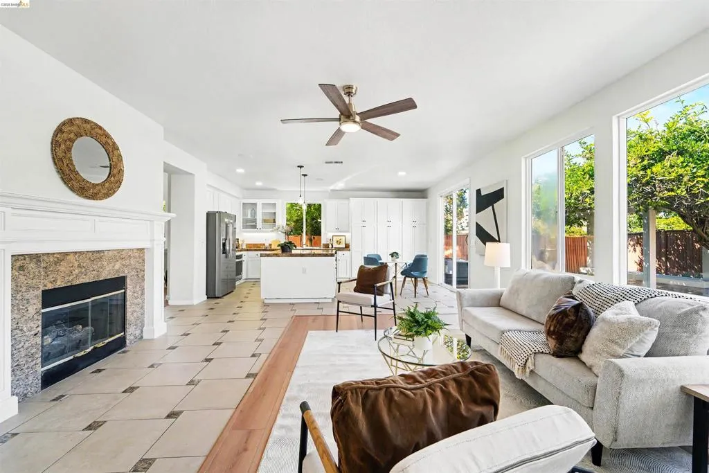 Bright open-concept living area featuring a fireplace with a white mantel and stone surround, large windows, ceiling fan, and a combination of tile and wood-finish flooring
