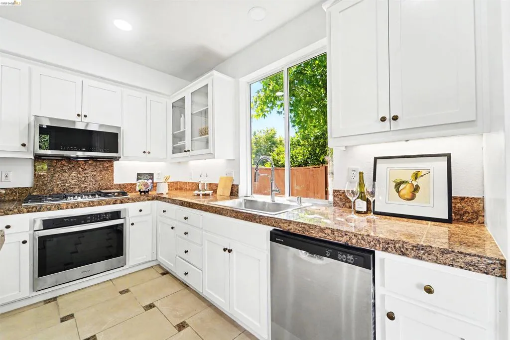 Contemporary kitchen featuring white cabinetry, stainless steel appliances, a window above the sink, stone countertops and backsplash, and tiled flooring