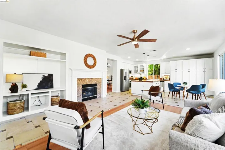 Open-concept living area featuring a stone fireplace with a white mantel, built-in shelving, a ceiling fan, and wood-finish flooring