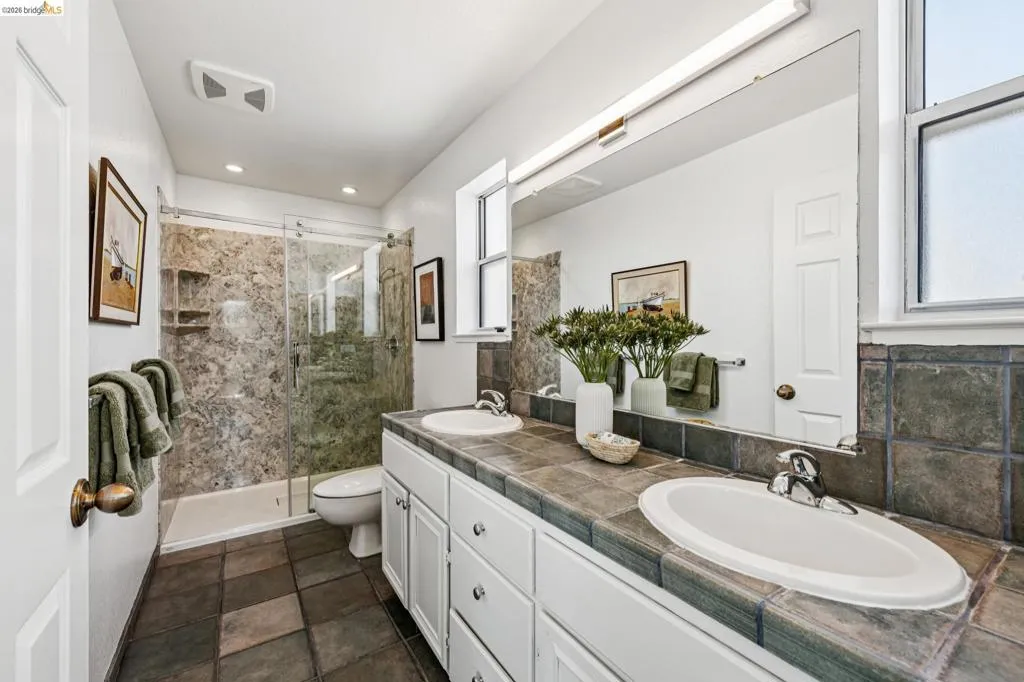 Bathroom featuring a dual vanity with tile countertop, tile flooring, and a glass-enclosed shower with built-in shelving