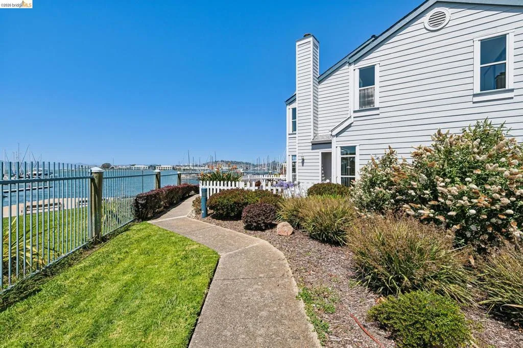 Waterfront property featuring a paved walkway, mature landscaping, and a white picket fence