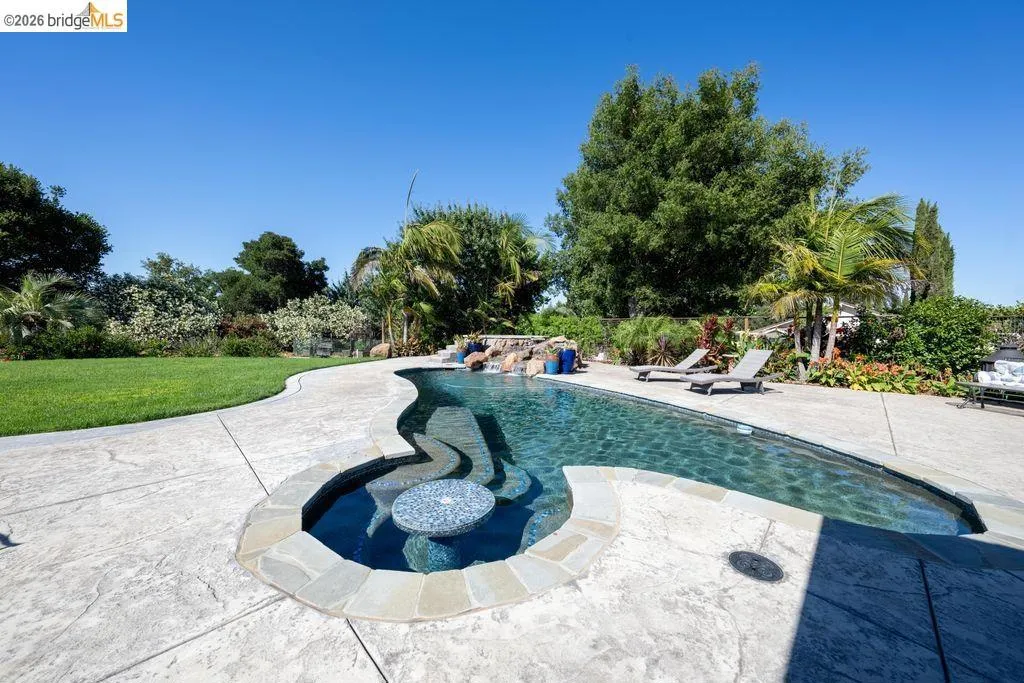 Curved swimming pool featuring a built-in spa with a mosaic table, rock waterfall, and surrounding stamped concrete patio