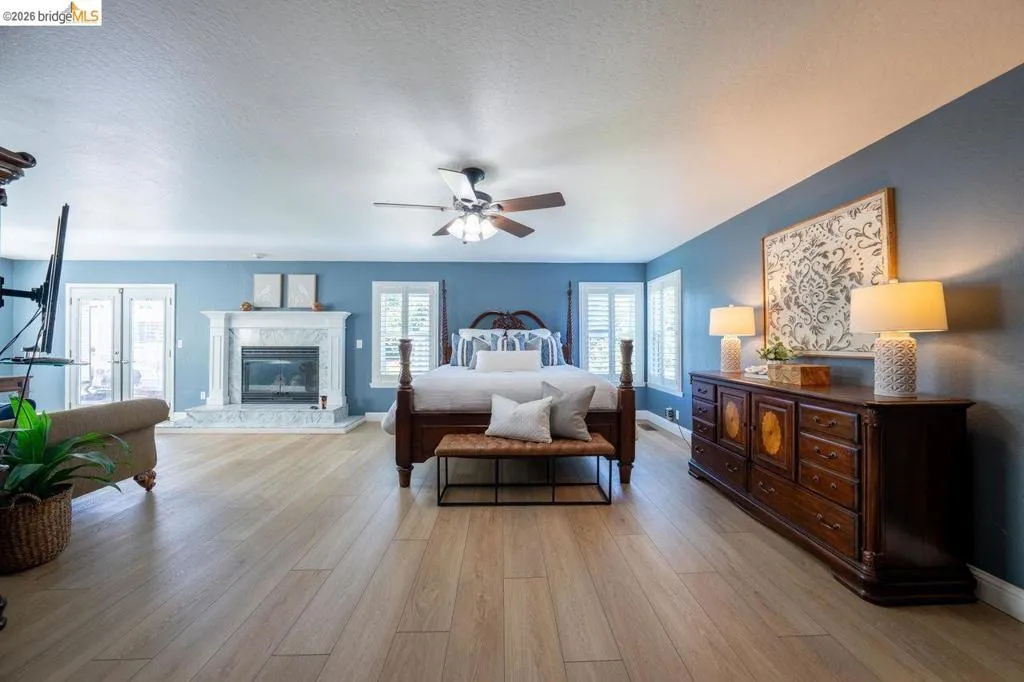 Spacious room featuring light wood-finish flooring, a ceiling fan, and a fireplace with a light-colored surround