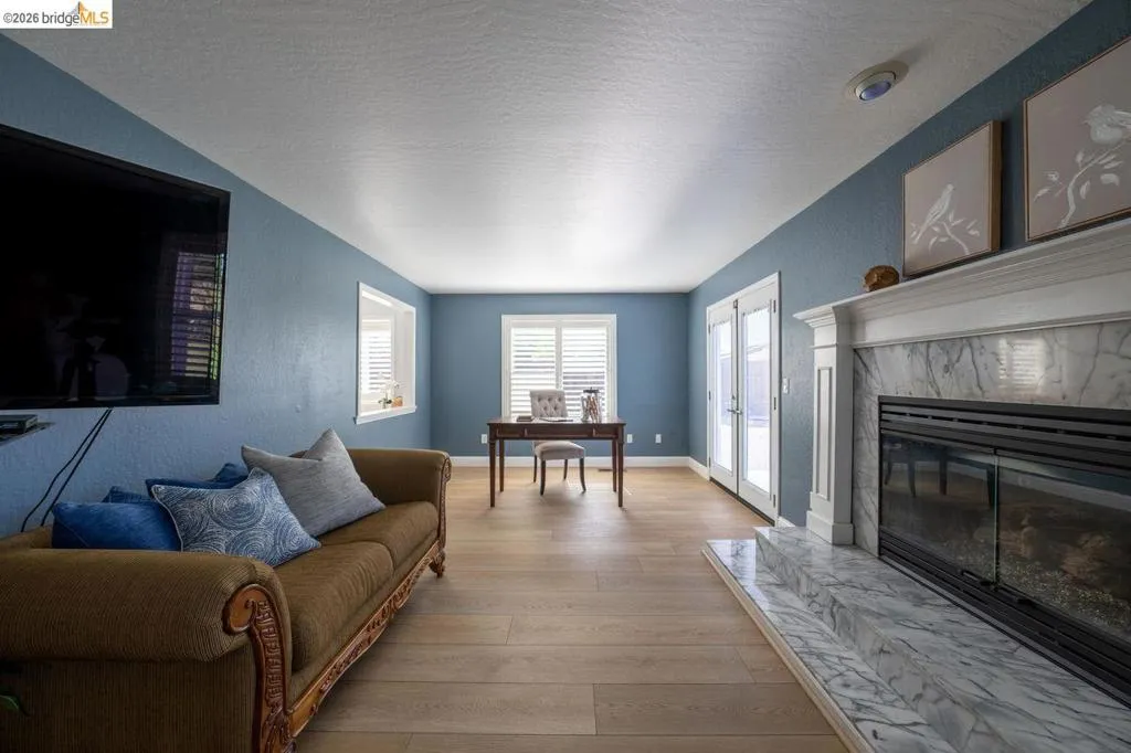 Living area with light wood-finish flooring, a fireplace featuring a white marble surround and hearth, and double French doors with white frames