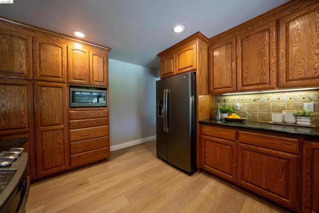 Kitchen featuring wood cabinetry, dark countertops, a built-in microwave, a stainless steel refrigerator, and wood-finish flooring