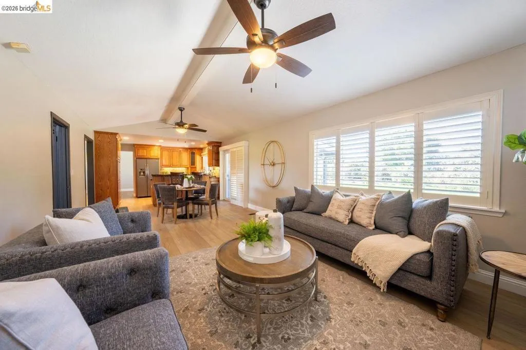 Open-concept living area with light wood-finish flooring, white walls, and vaulted ceilings featuring exposed beams