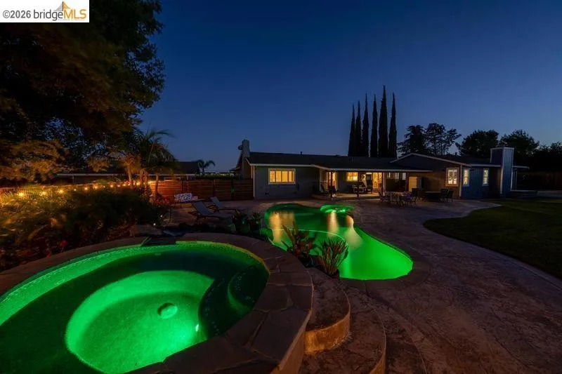 Resort-style backyard featuring a brightly lit swimming pool, an illuminated spa, an expansive concrete patio, and mature landscaping