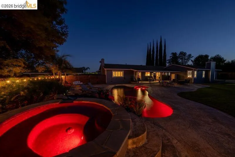 Rear exterior featuring a freeform swimming pool with red lighting, an attached spa, a concrete patio, and a manicured lawn