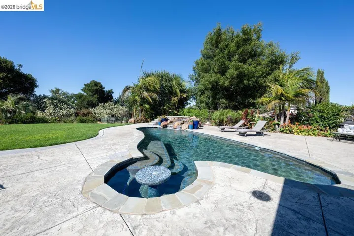 Curved swimming pool featuring a built-in spa with a mosaic table, rock waterfall, and surrounding stamped concrete patio