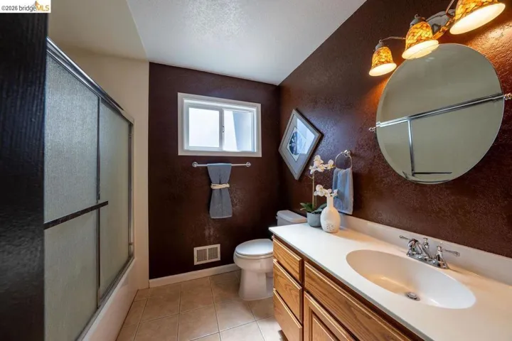 Bathroom featuring a wood-finish vanity with an integrated sink, a large oval mirror, and a shower-tub combination with sliding frosted glass doors
