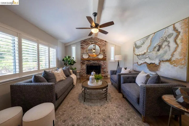 Vaulted ceiling living area featuring a brick fireplace with wood mantel, large windows with white louvered shutters, ceiling fan, and wood-finish flooring