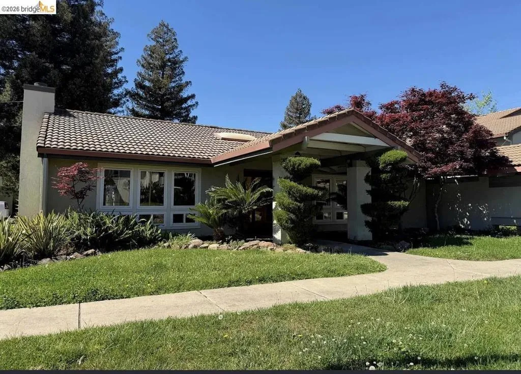Exterior featuring a tile roof, light green stucco siding, a prominent chimney, a large picture window, and a covered entry with manicured shrubbery