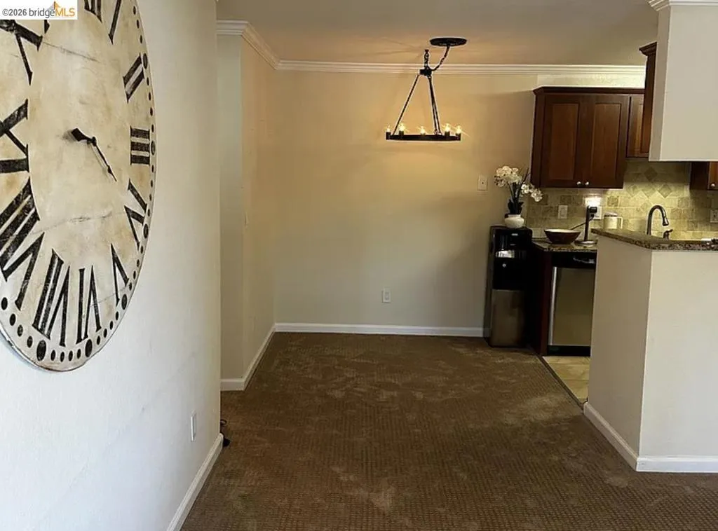 Dining area featuring a chandelier-style light fixture, crown molding, and carpet flooring, with a partial view of a kitchen with dark wood-finish cabinetry, tile backsplash, and a built-in sink