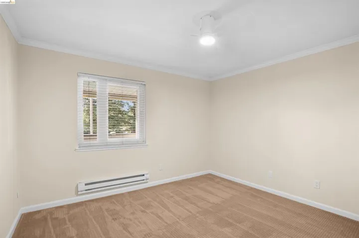 Carpeted interior space featuring a window with horizontal blinds, wall-mounted baseboard heating unit, ceiling-mounted light fixture, and white crown molding
