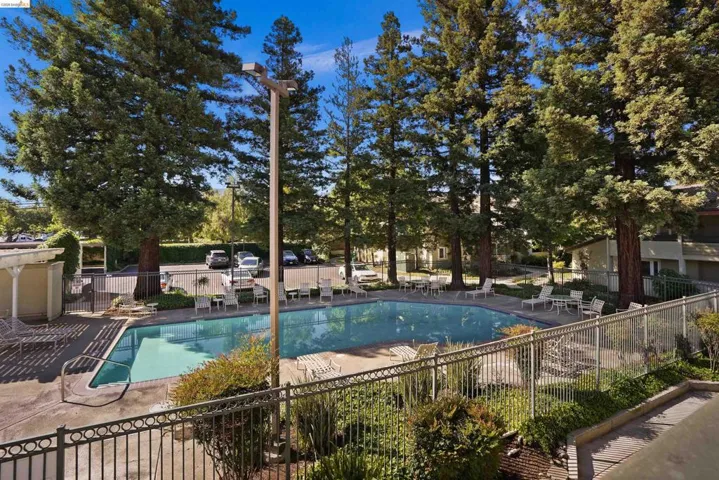 Community swimming pool surrounded by mature trees, featuring a paved deck, perimeter fencing, and seating areas