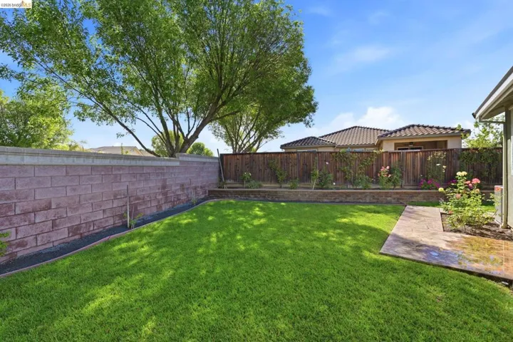 Expansive backyard featuring a lush lawn, a decorative block retaining wall, mature trees, and a stamped concrete patio area