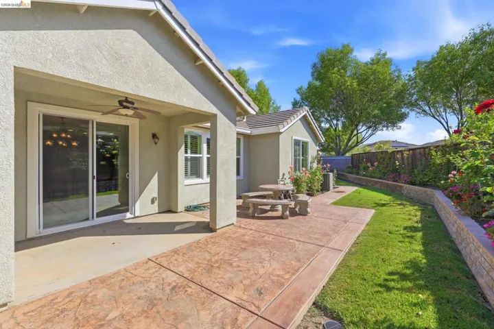 Expansive backyard featuring a stamped concrete patio, covered outdoor living area with ceiling fan, turf lawn, and a stone retaining wall with mature plantings