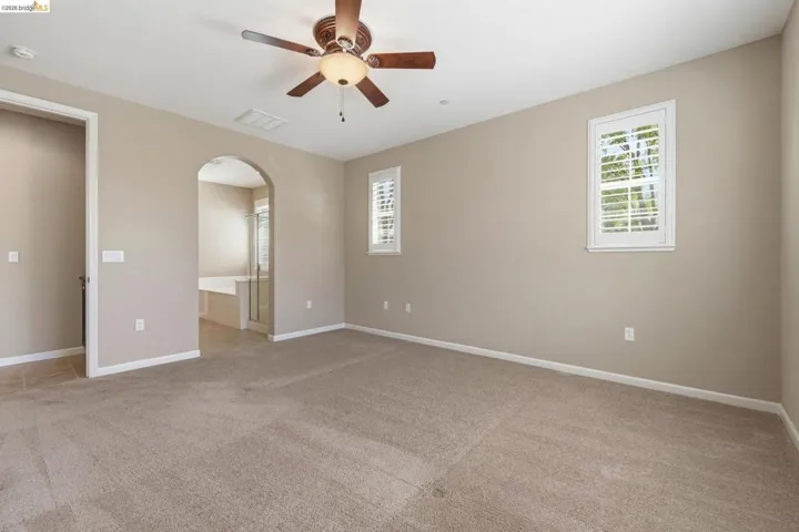 Carpeted room featuring a ceiling fan with integrated lighting, two windows with white plantation shutters, and an arched entry leading to a tiled bathroom with a built-in tub and glass-enclosed shower