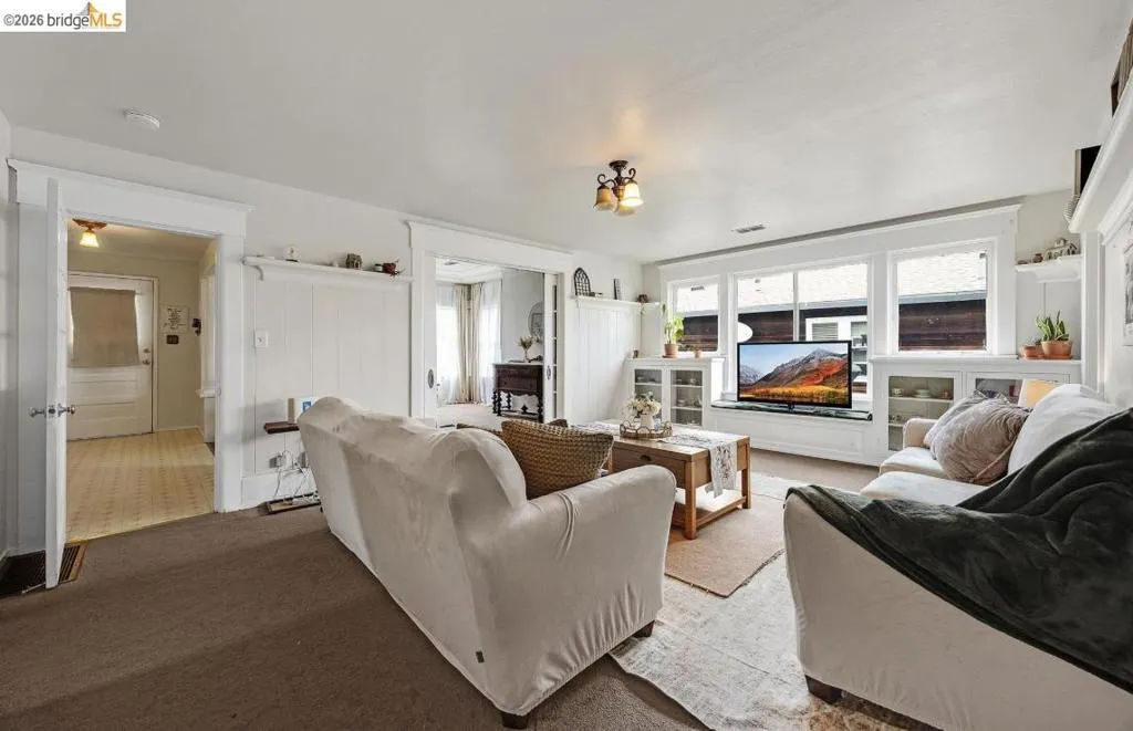 Living area featuring white paneled walls, a wall of built-in cabinetry with glass-front display cases, and window trim