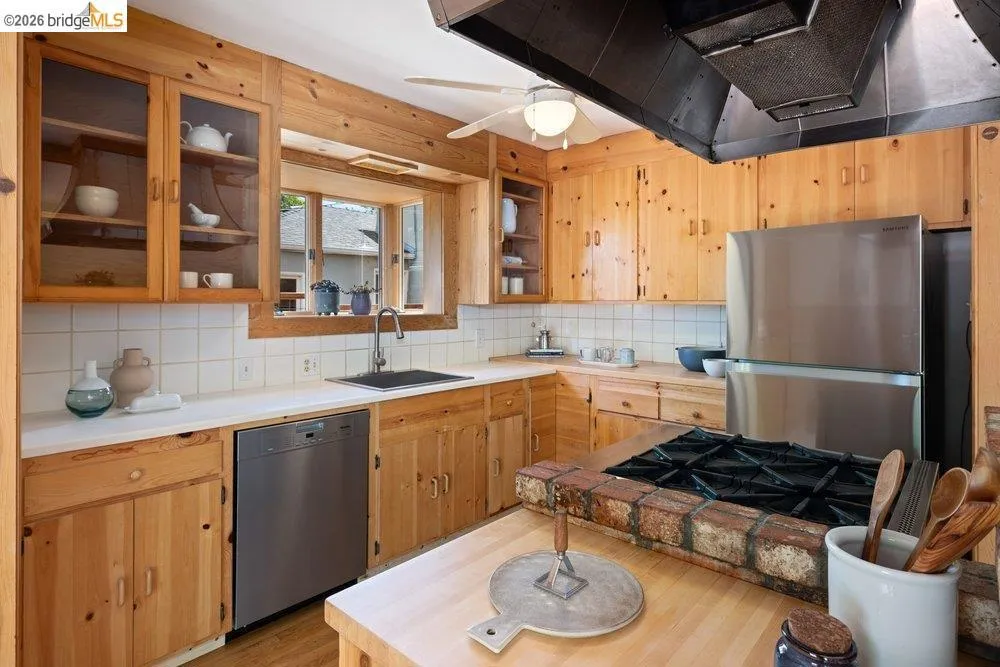Kitchen featuring natural wood cabinetry, white countertops, tile backsplash, stainless steel appliances, and a built-in cooktop with a brick surround