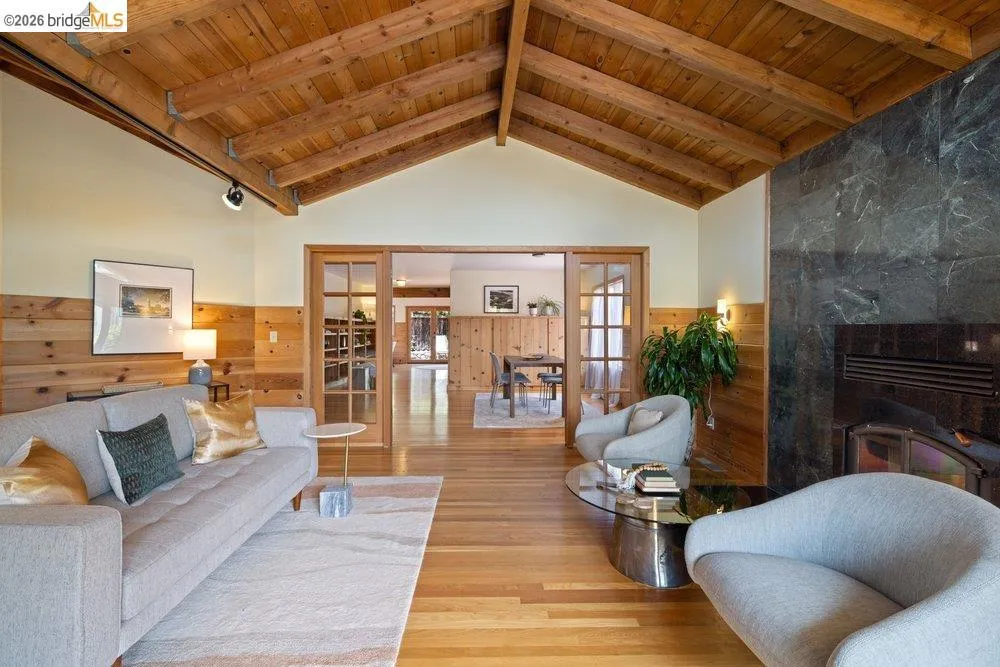 Vaulted wood-plank ceiling with exposed beams, hardwood flooring, full-height stone fireplace surround, wood paneling, and glass-paned sliding doors
