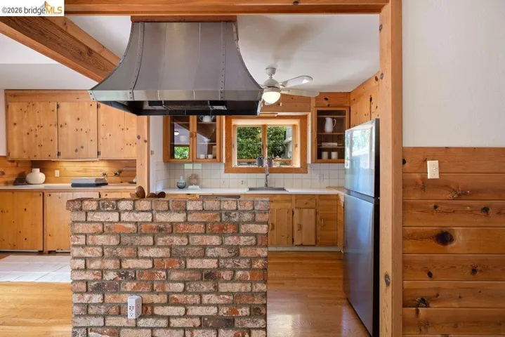 Kitchen featuring a brick half-wall, wood-finish cabinetry, stainless steel range hood, white countertops, and light wood-finish flooring