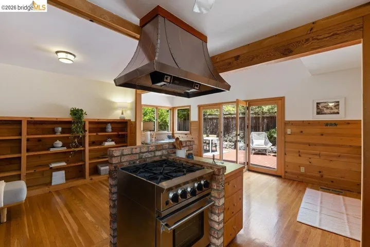 Stainless steel range with a custom metal hood, brick island base, wood-finish flooring, and exposed wood ceiling beams