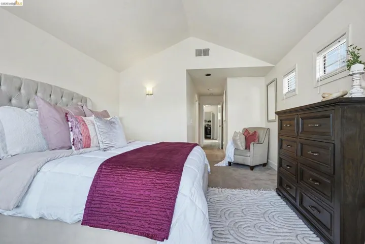 Vaulted ceilings enhance this carpeted bedroom featuring multiple windows, a wall sconce, and a hallway entry