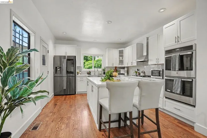 Open-concept kitchen featuring an oversized island with seating, white shaker cabinetry, stainless steel appliances, and wood-finish flooring