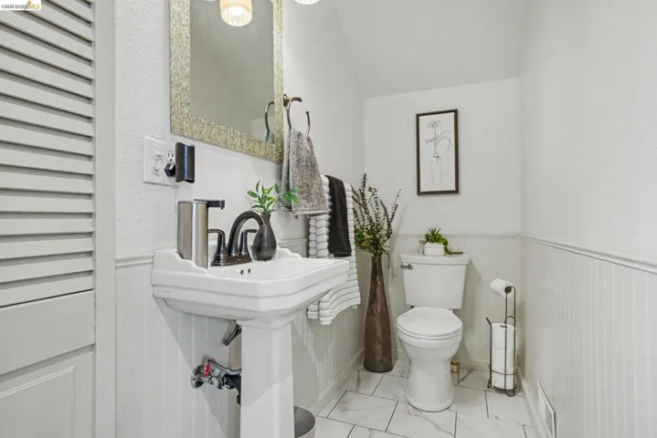 Pedestal sink with oil-rubbed bronze faucet and hardware, white beadboard wainscoting, white toilet, white tile flooring with gray veining, and a decorative framed mirror
