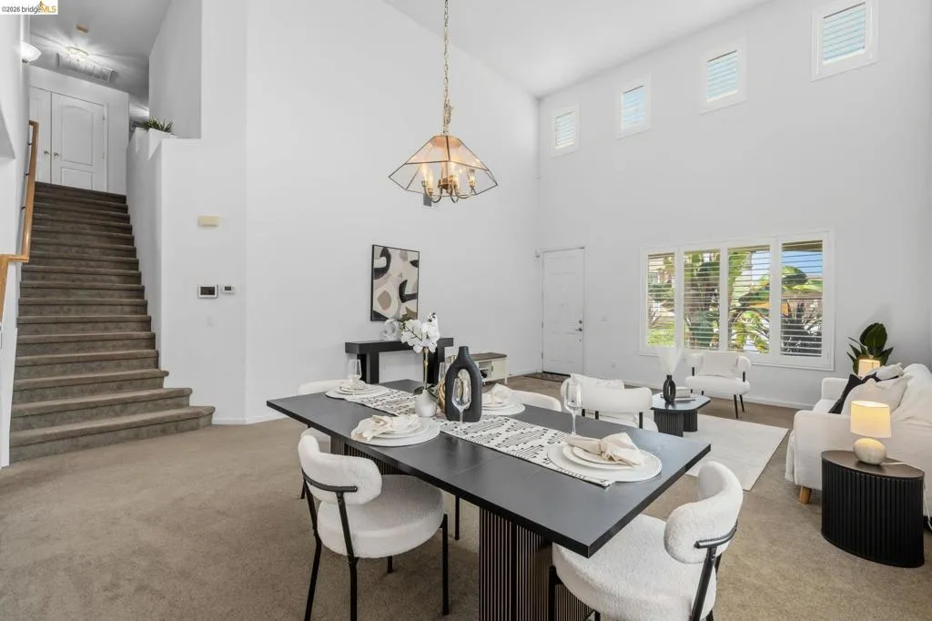Two-story living area with vaulted ceilings, large windows with plantation shutters, a prominent staircase with carpeted treads and wood railings, and a decorative chandelier